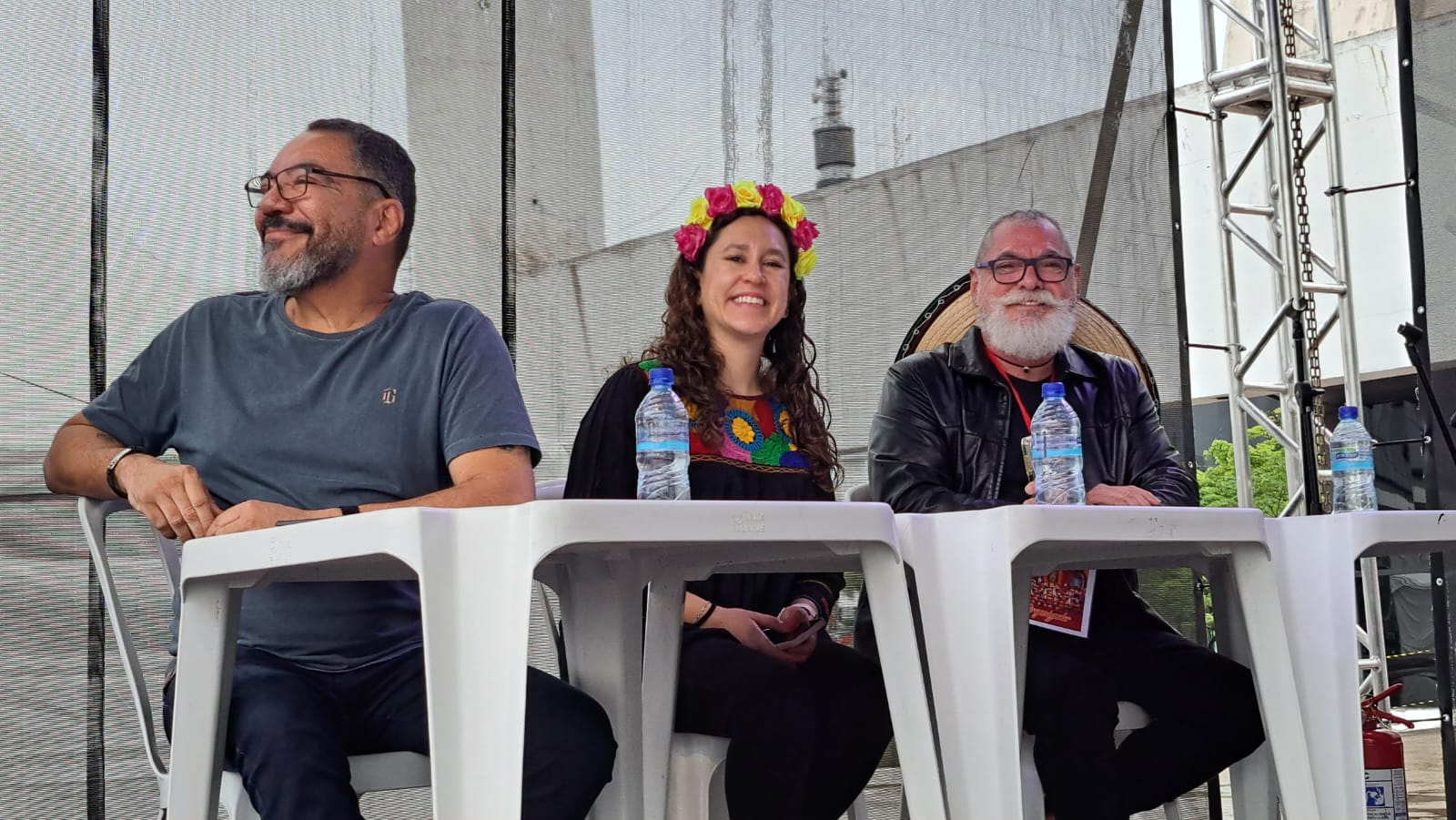 Três integrantes de mesa de avaliação permanecem sentados sob estrutura de evento ao ar livre