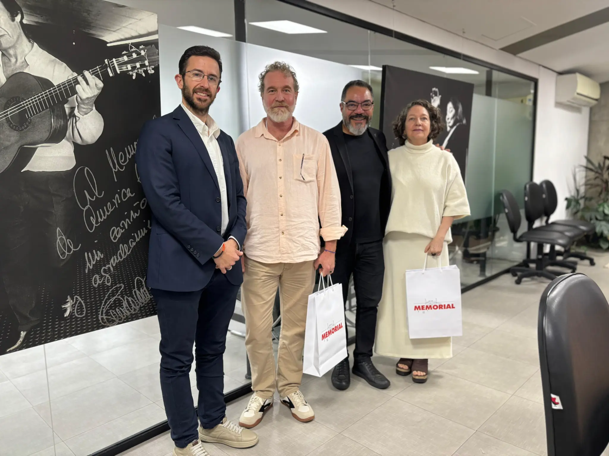 Quatro participantes de agenda cultural posam com sacolas do Memorial diante de painéis expositivos