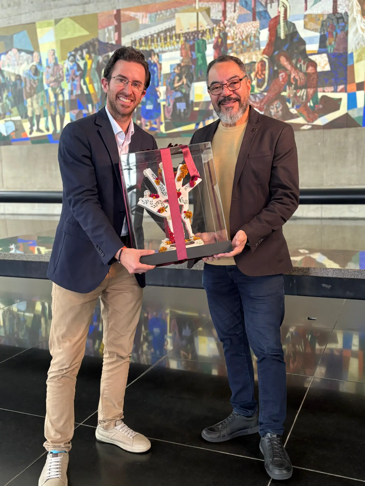 Rodrigo Vázquez e João Carlos Corrêa seguram uma caixa transparente com escultura em forma de mão diante de mural colorido