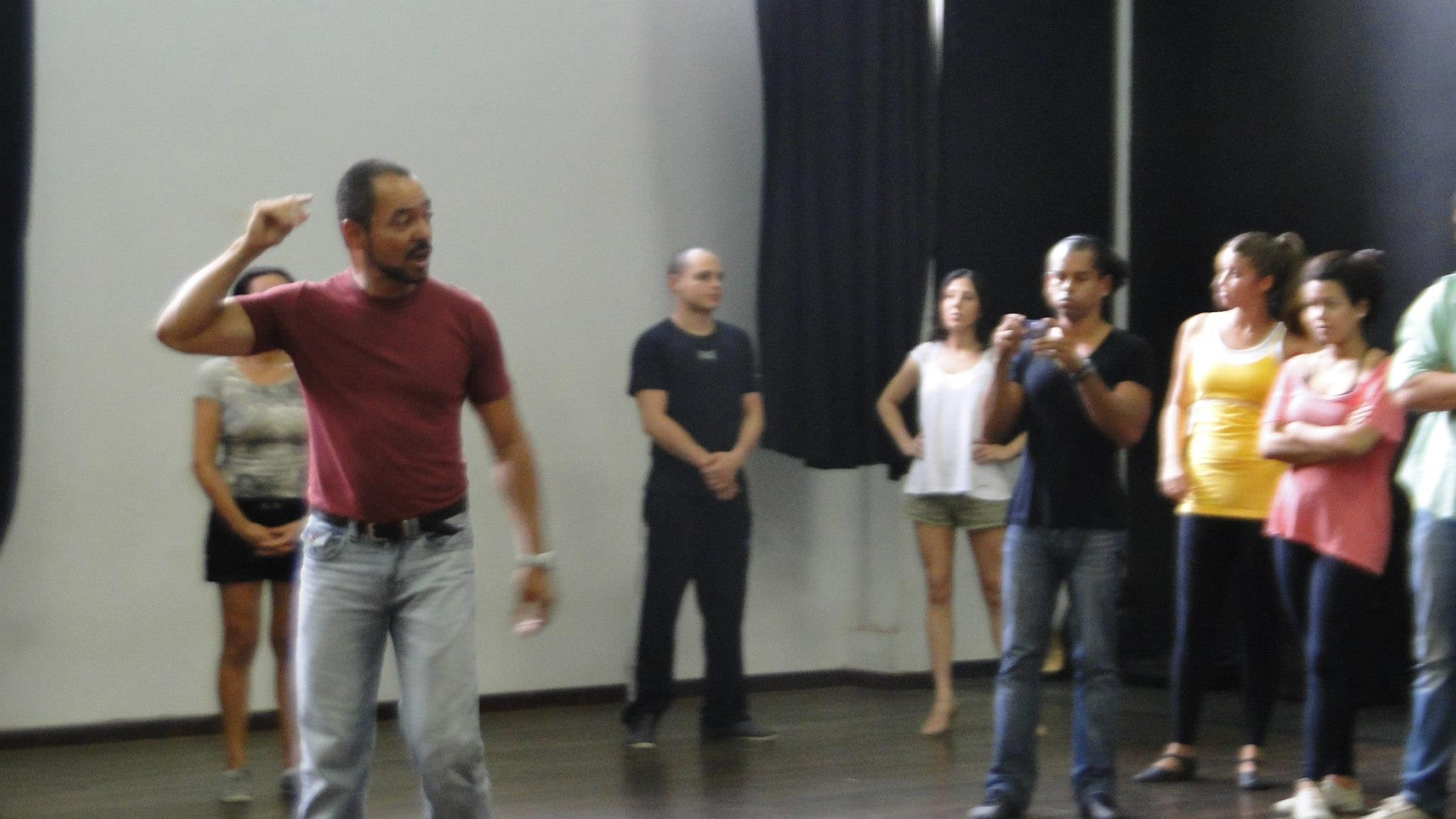 Homem de camiseta vermelha gesticula em primeiro plano enquanto participantes observam em uma sala de dança.