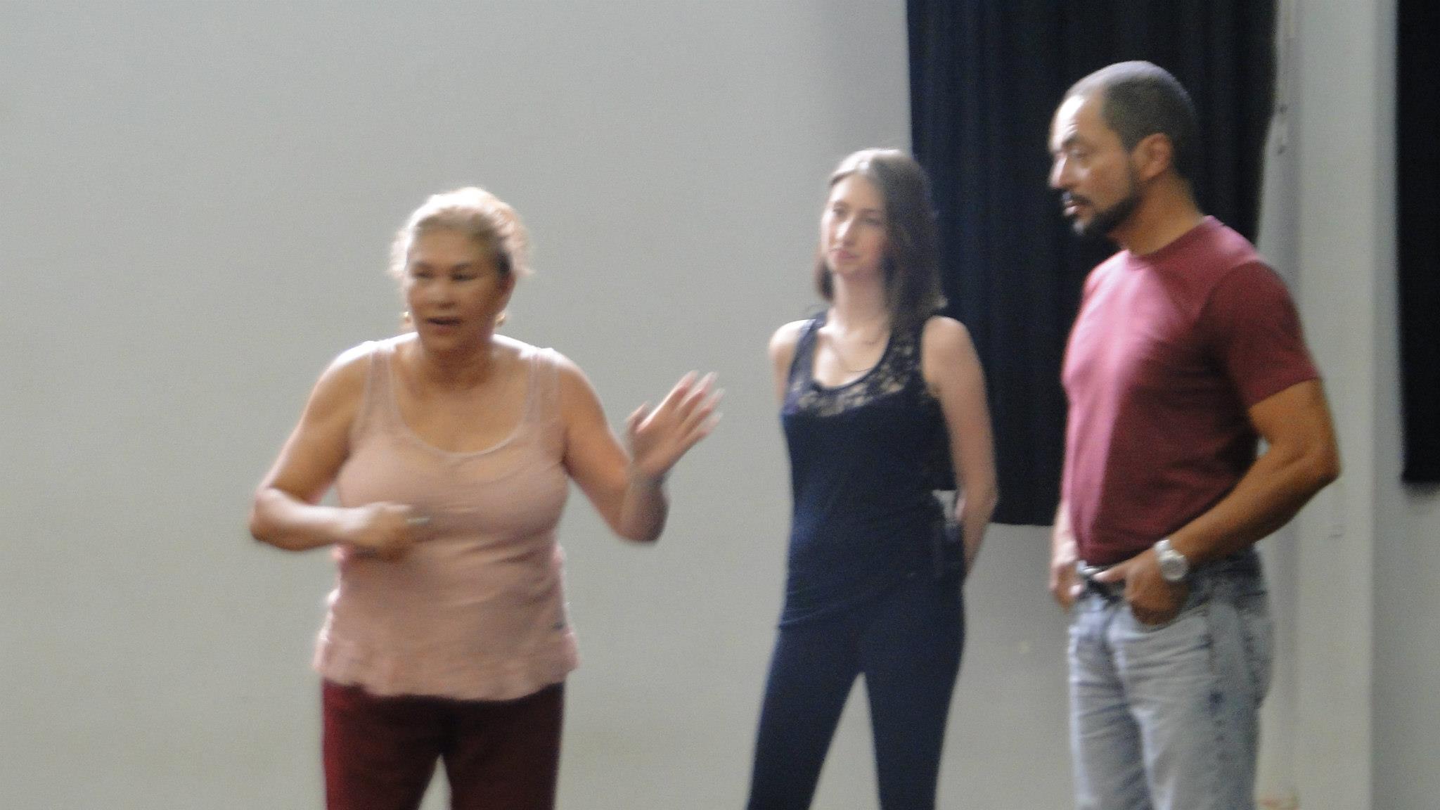 Mulher de blusa rosa gesticula durante uma explicação, acompanhada por duas pessoas em uma sala de dança.