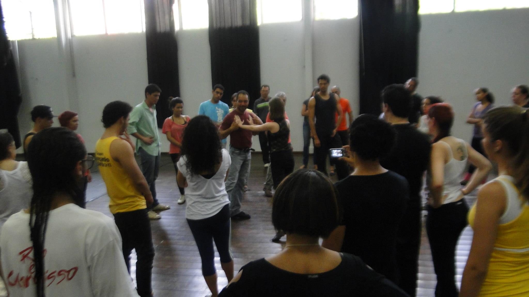 Casal demonstra movimentos no centro da sala enquanto participantes observam ao redor.