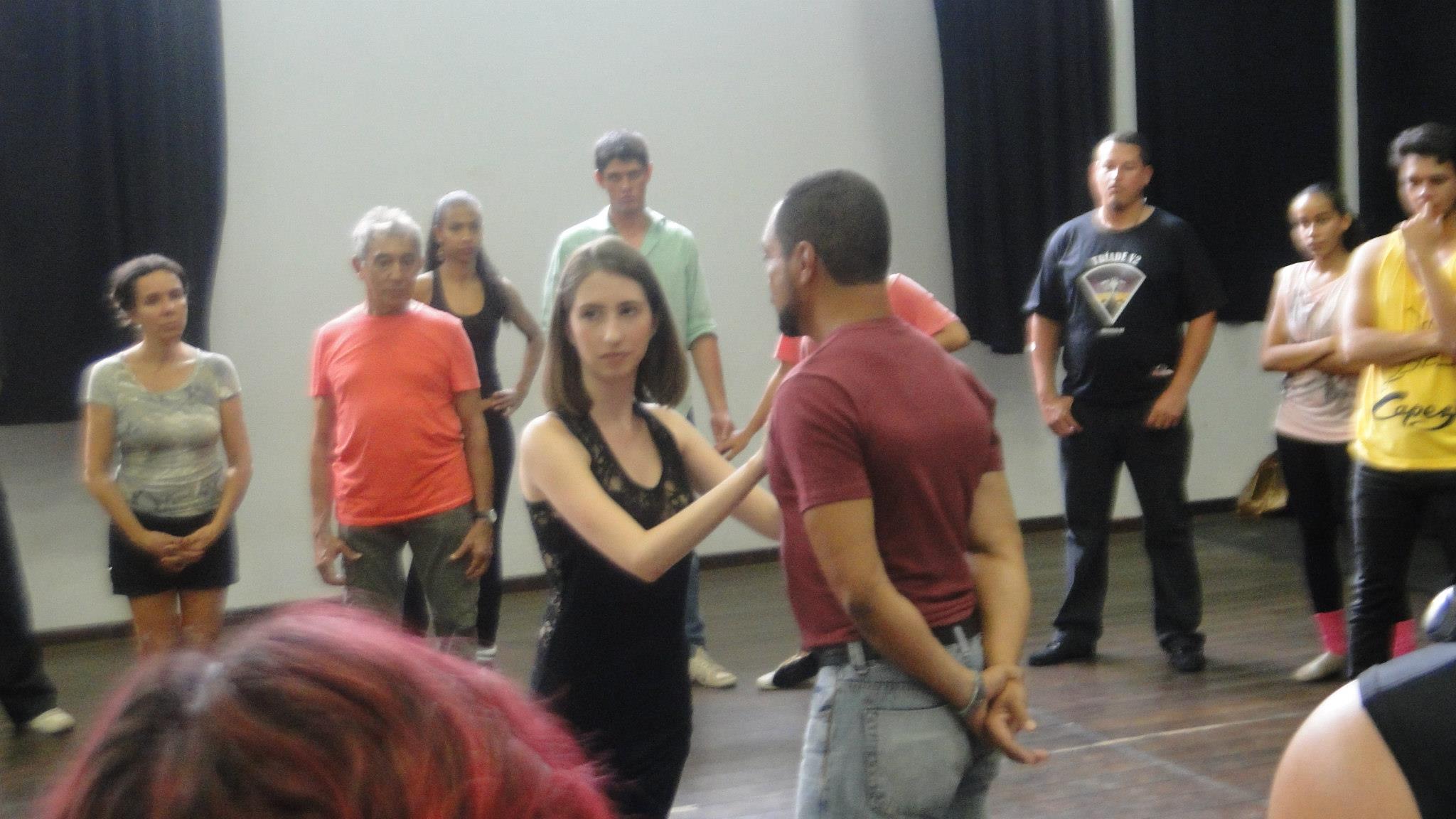 Casal dança no centro do estúdio cercado por participantes que acompanham a demonstração.