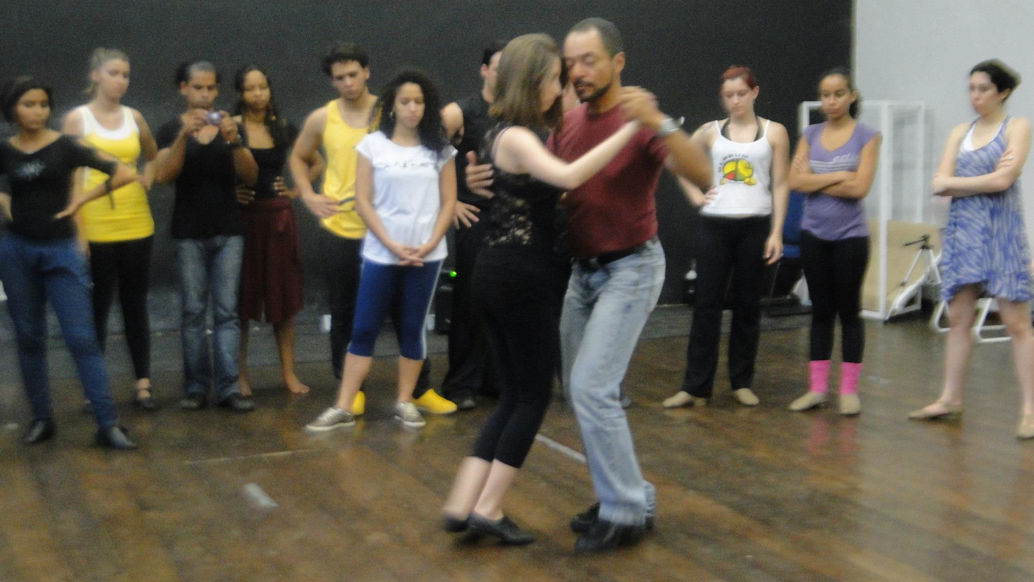 Casal pratica tango no centro do salão enquanto participantes observam em pé ao redor.