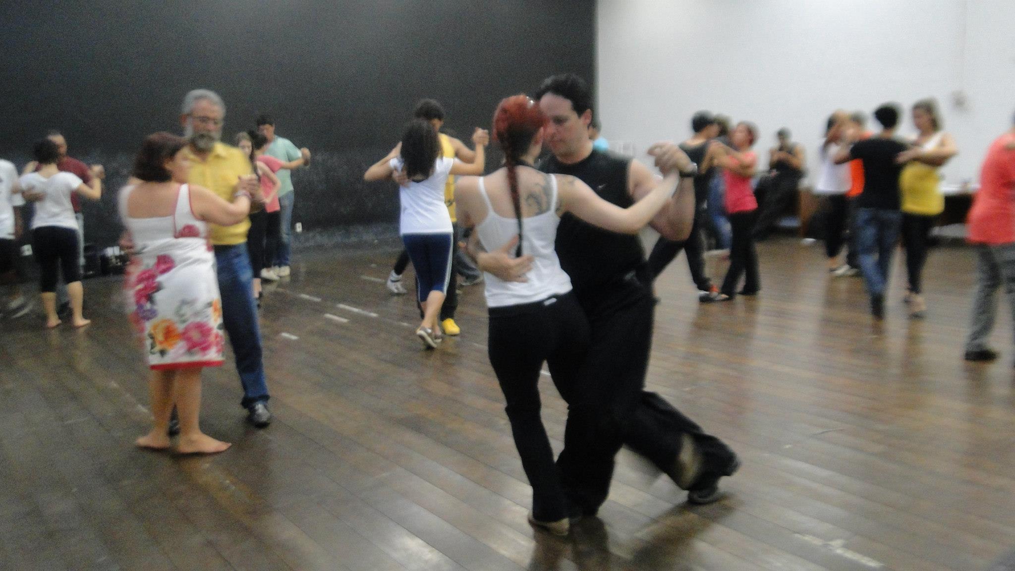 Várias duplas praticam passos de tango em um salão amplo com piso de madeira.