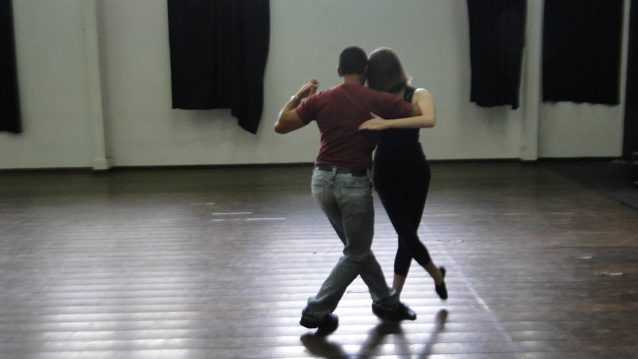 Casal visto de costas pratica tango no centro de um salão com piso de madeira.
