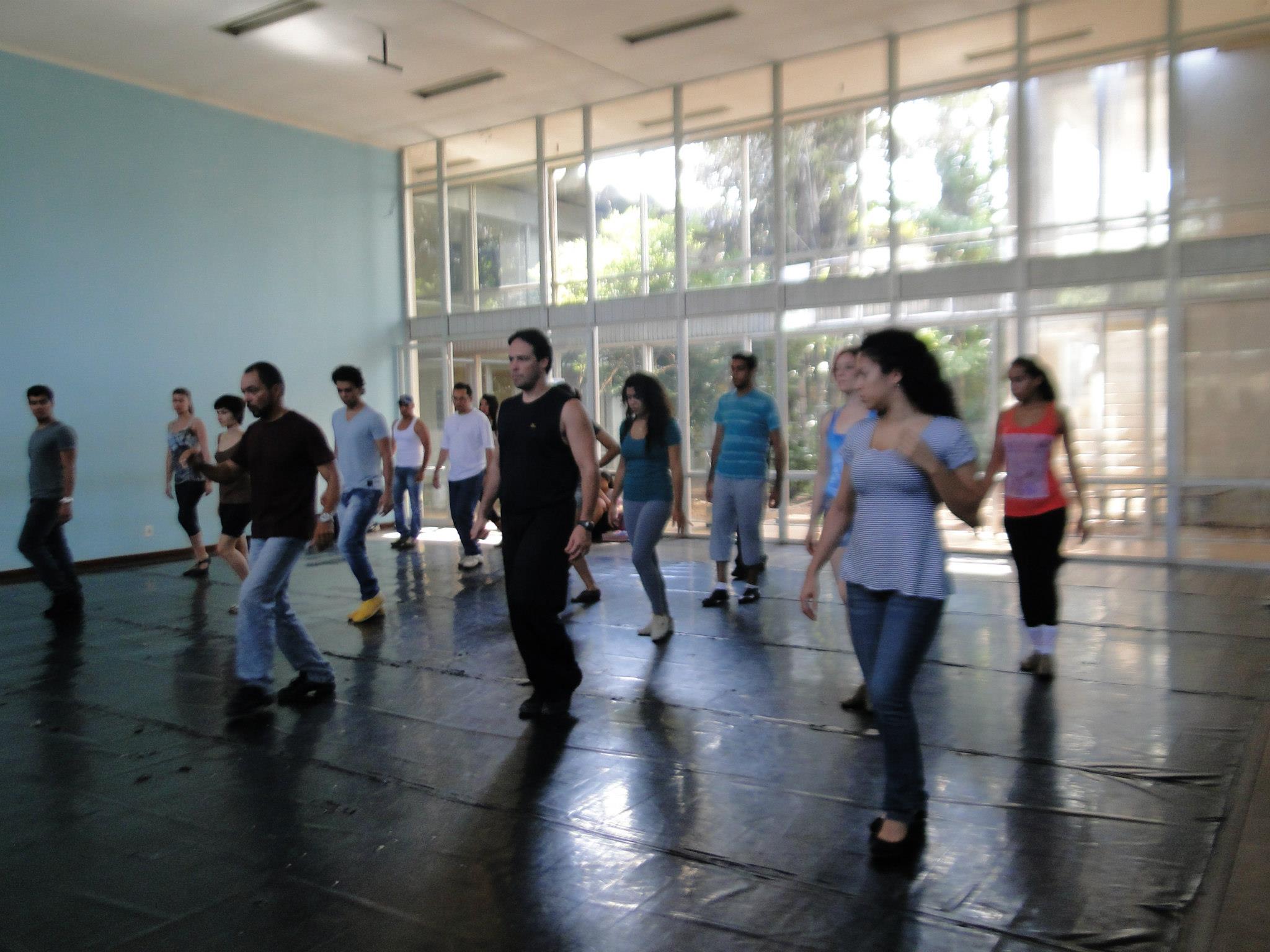 Grupo de participantes pratica passos em uma sala ampla com piso escuro e janelas de vidro.