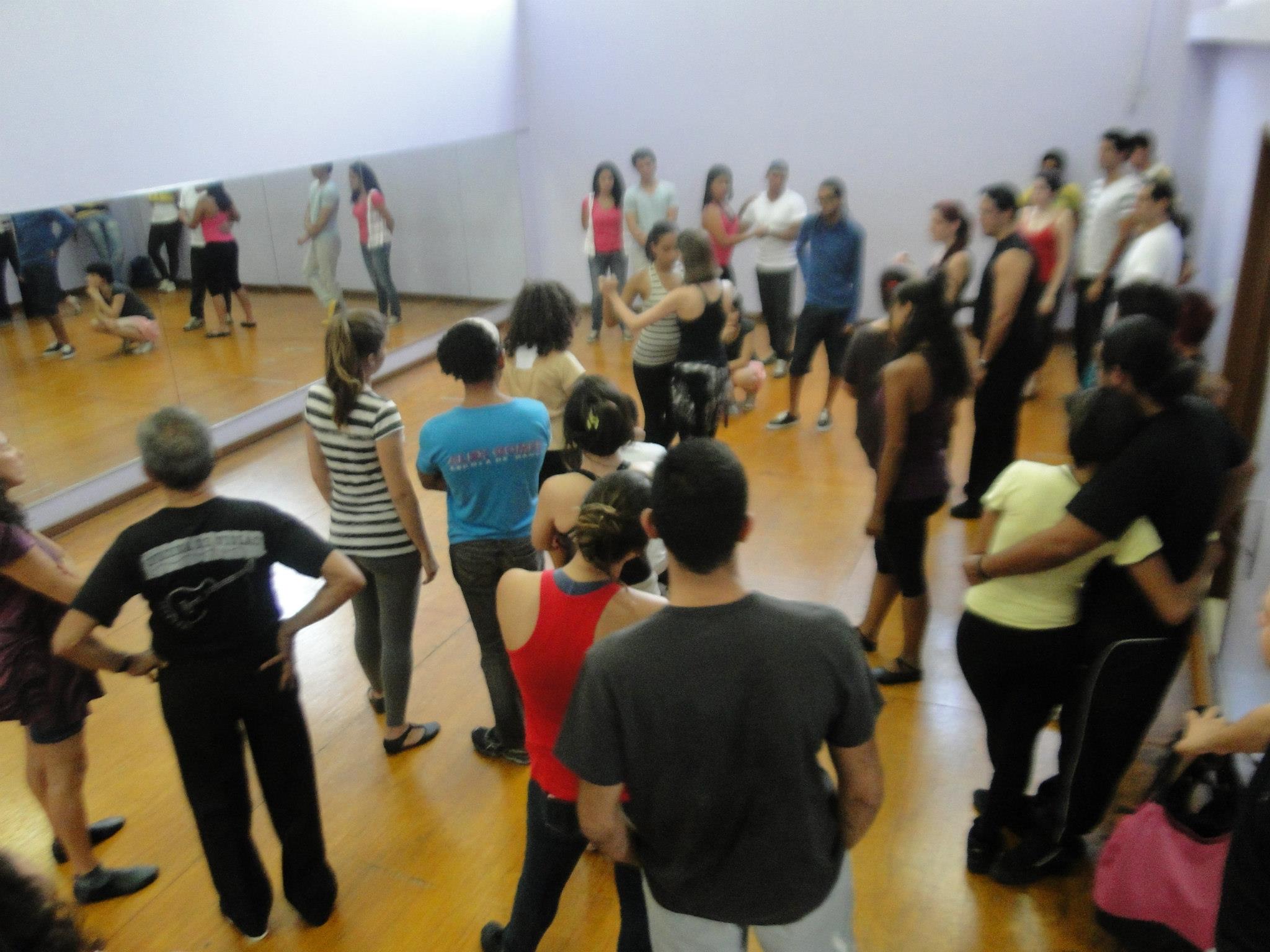 Participantes formam pares em um estúdio de dança com piso de madeira e parede espelhada.