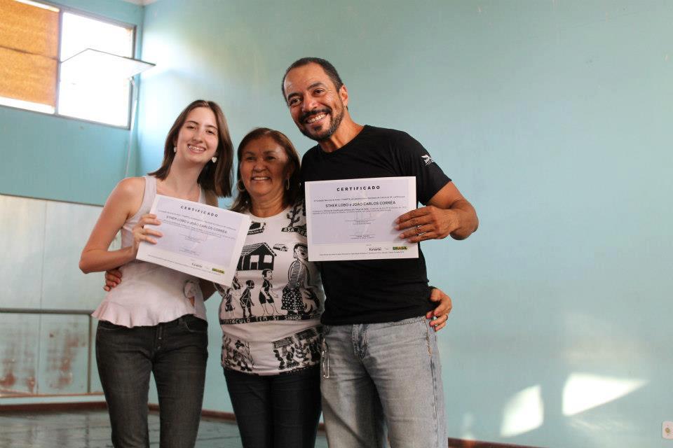 Três pessoas posam abraçadas, com duas delas segurando certificados em uma sala de paredes azuis.