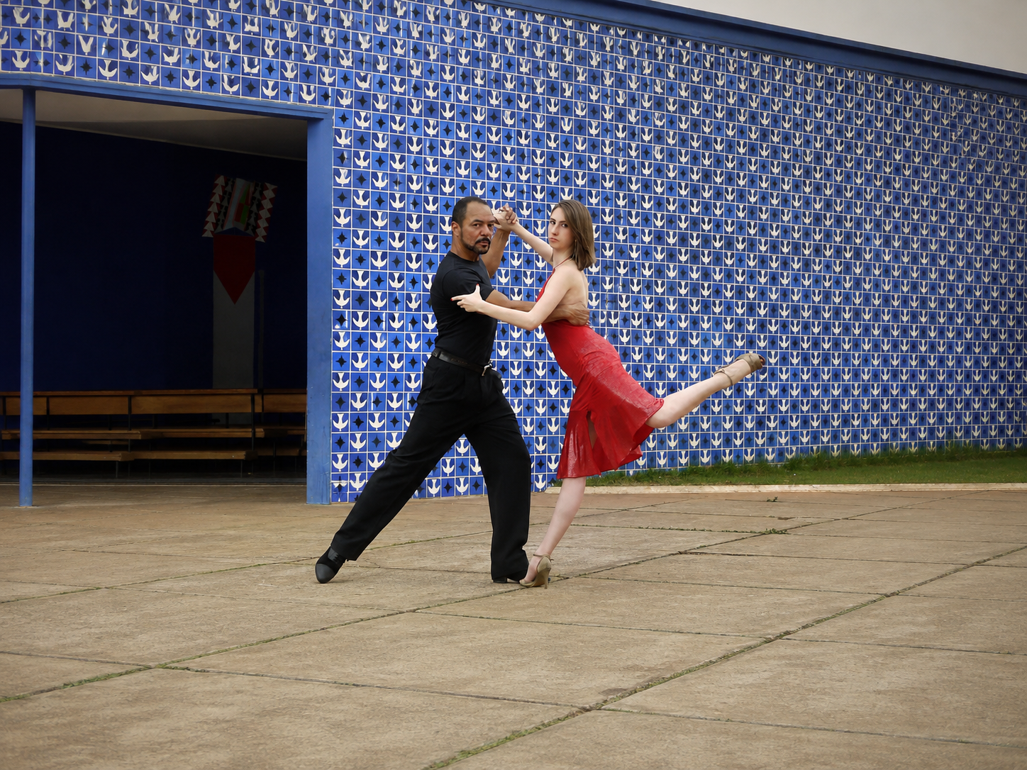 João Carlos Corrêa e uma parceira dançando tango em plano aberto, diante de painel de azulejos e área externa de equipamento cultural
