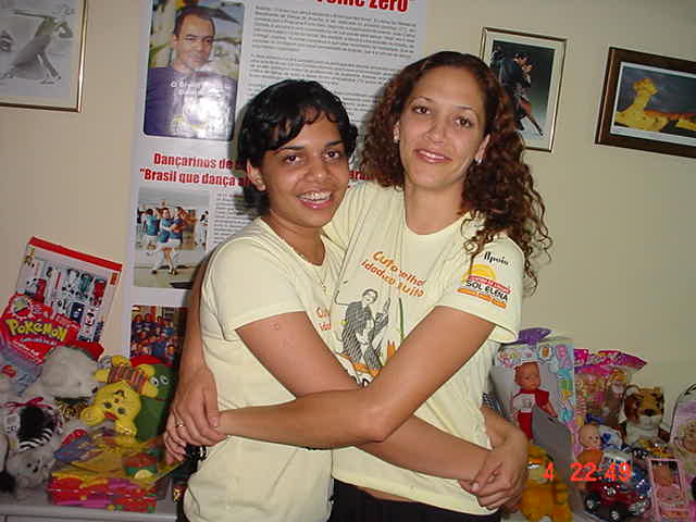 Duas integrantes com camisetas amarelas da programação abraçadas diante dos brinquedos arrecadados