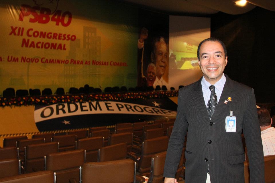 João Carlos Corrêa posa em auditório diante de palco e painel de congresso.