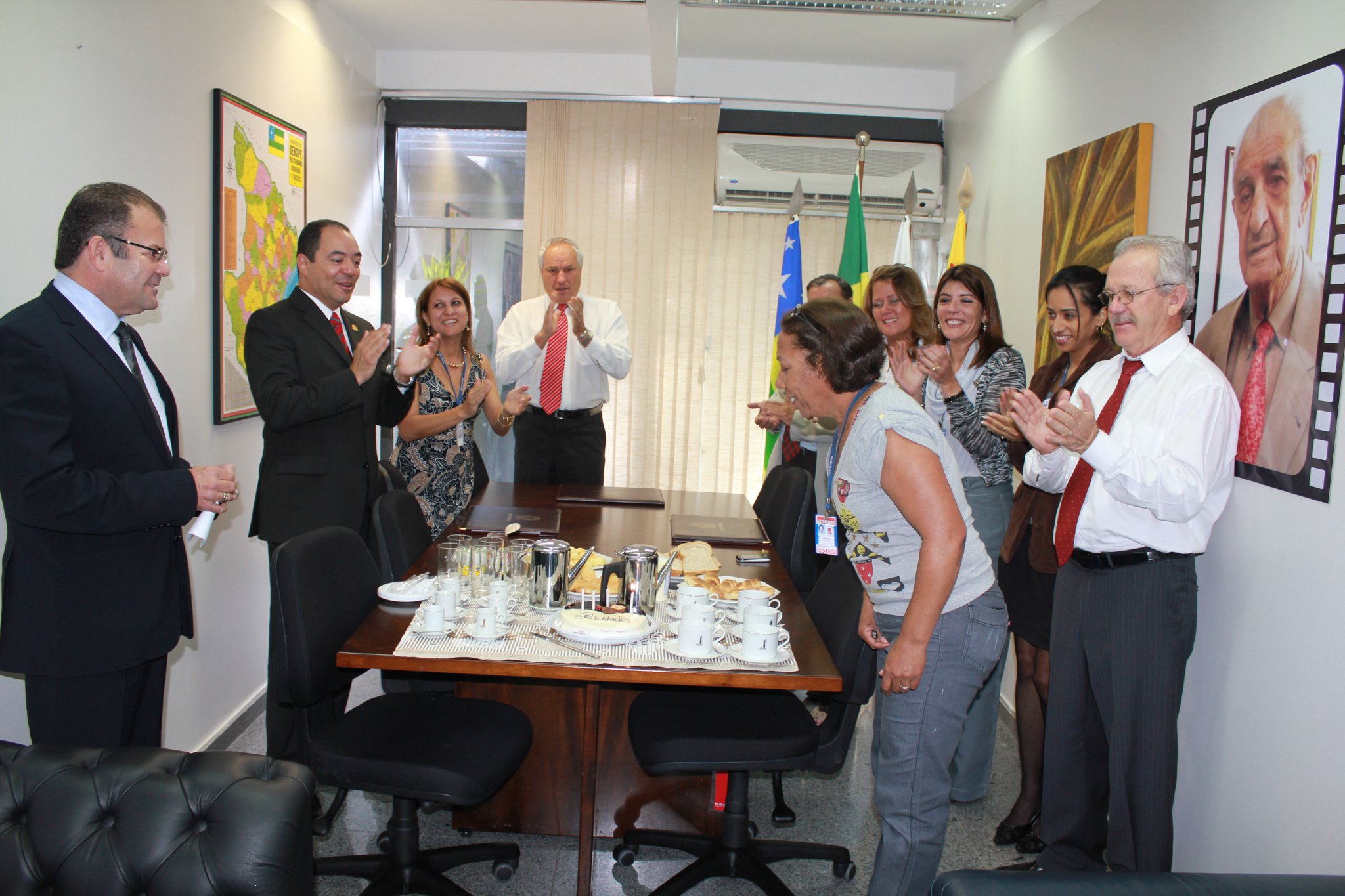 Grupo reunido em sala de trabalho aplaude ao redor de uma mesa com bolo, xícaras e garrafas térmicas.