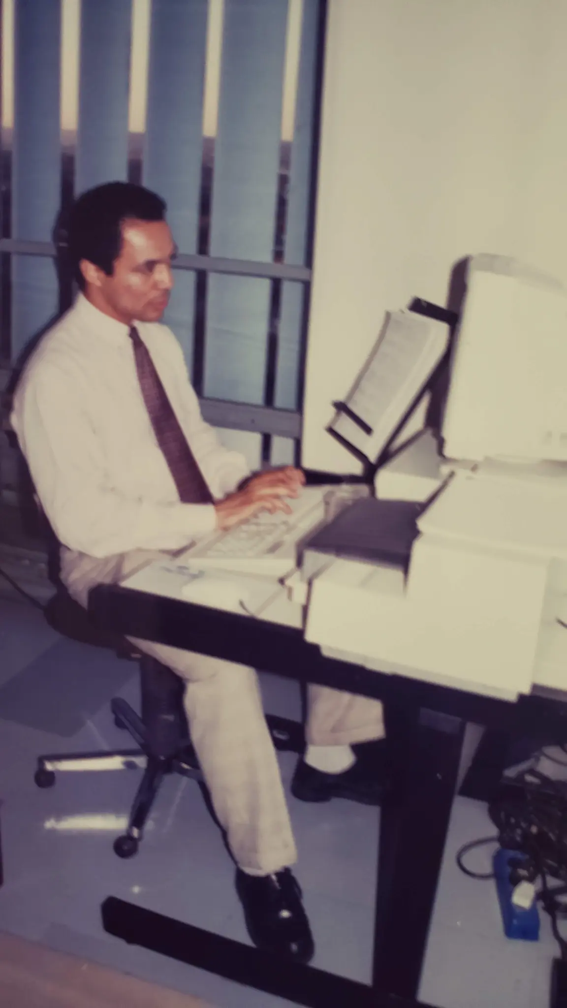 João Carlos Corrêa sentado diante de computador com monitor de tubo e suporte para documentos em ambiente de escritório.