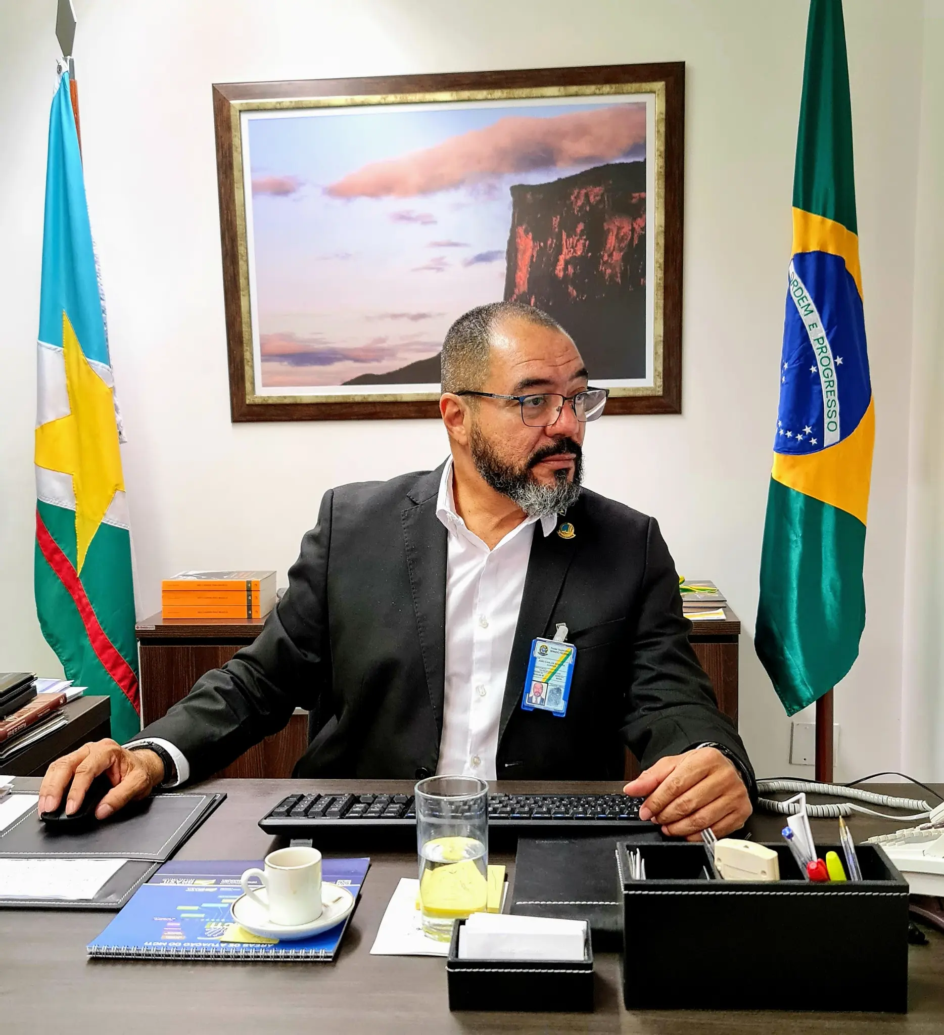 João Carlos Corrêa sentado à mesa com computador, documentos e bandeiras institucionais ao fundo.