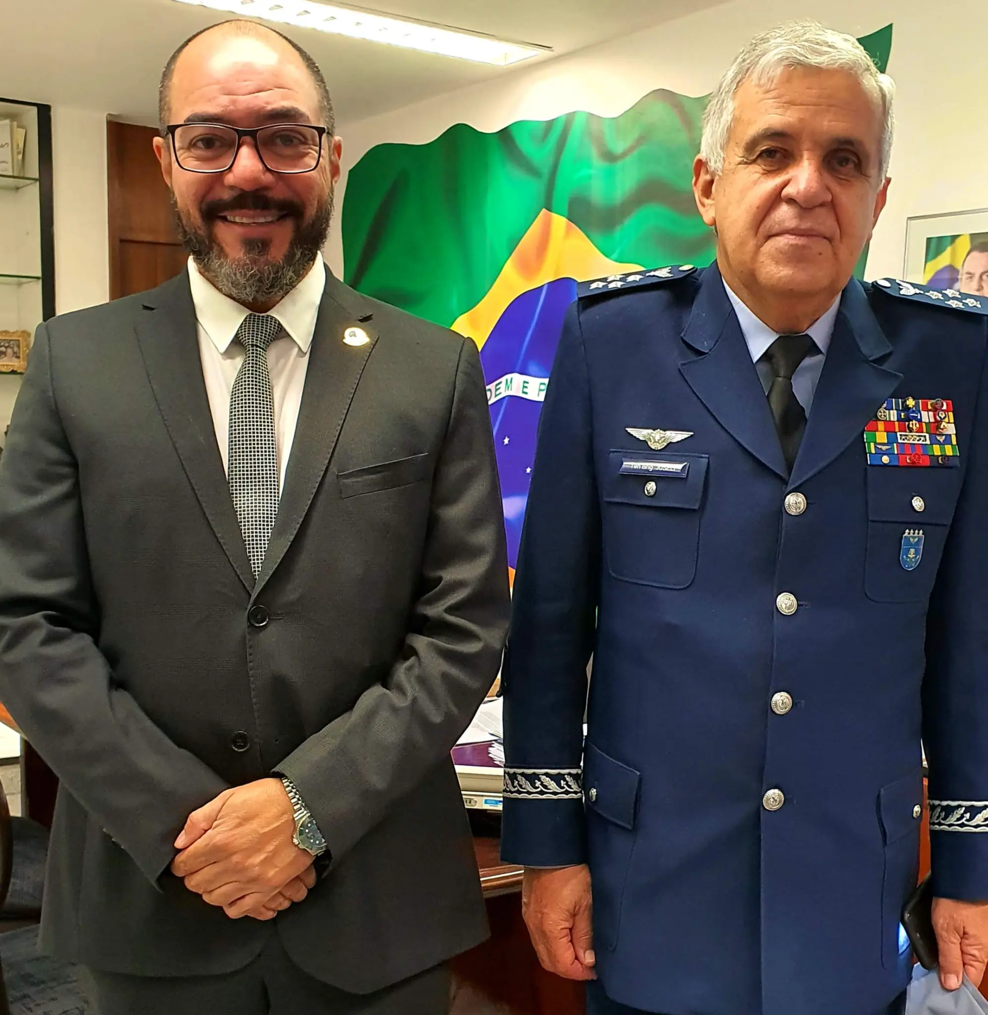 João Carlos Corrêa e um homem de uniforme militar posam diante de uma bandeira do Brasil em ambiente de gabinete.