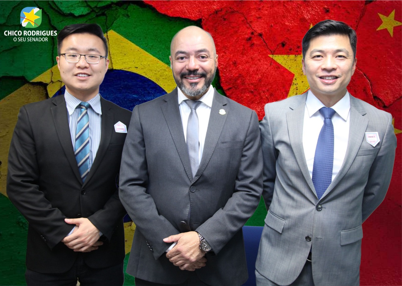 João Carlos Corrêa entre dois visitantes diante de composição visual com as bandeiras do Brasil e da China.