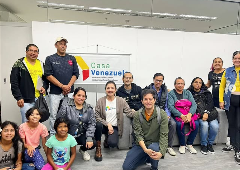 Un grupo de adultos y niños posa en un espacio interior frente a un banner con el nombre y el logotipo de Casa Venezuela