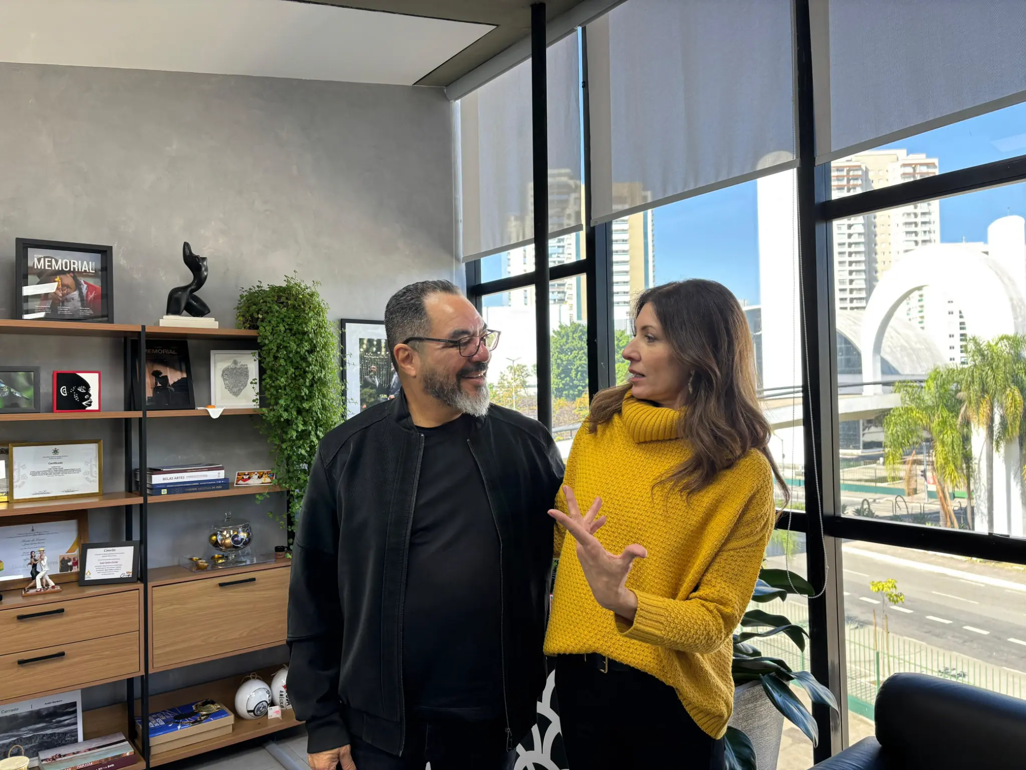 João Carlos Corrêa y Blanca Montilla conversan de pie en un entorno de trabajo con una estantería y vista al conjunto arquitectónico del Memorial