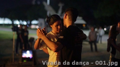 Casal dança de mãos dadas ao ar livre durante a programação noturna, com outras pessoas ao fundo.