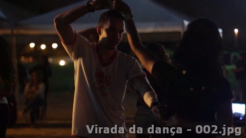 Casal dança em uma área externa à noite, com iluminação suave ao fundo.