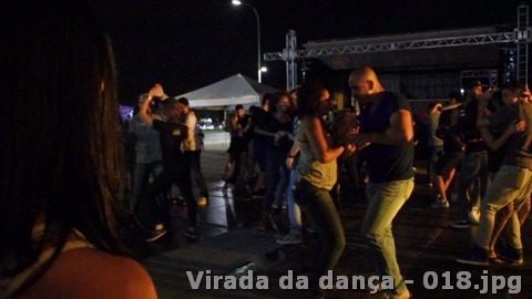 Várias pessoas dançam em pares em espaço aberto à noite, com palco e tendas ao fundo.