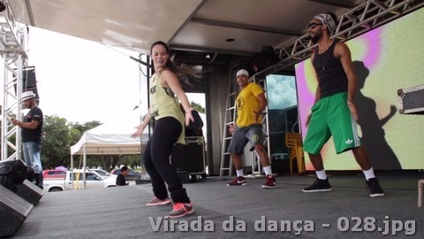 Três participantes realizam uma coreografia em palco coberto com telão ao fundo.