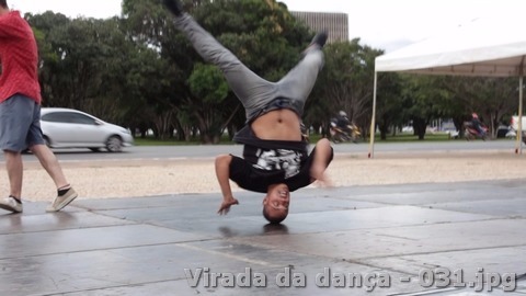 Dançarino realiza movimento acrobático apoiado na cabeça sobre piso ao ar livre.