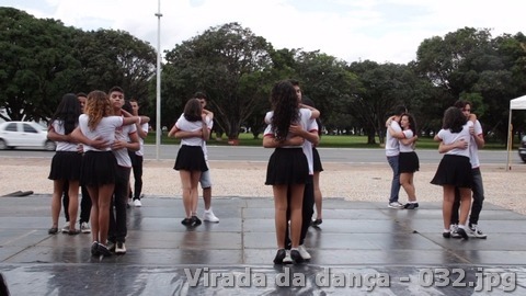 Jovens dançam em pares sobre uma plataforma ao ar livre, com árvores e via pública ao fundo.