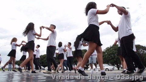 Grupo jovem dança em pares sobre uma plataforma ao ar livre usando roupas claras e escuras.