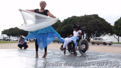 Dançarina movimenta um tecido branco ao lado de uma pessoa em cadeira de rodas durante apresentação ao ar livre.