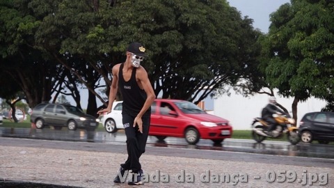 Pessoa com roupa preta, boné e máscara prateada realiza movimento de dança próximo a uma via pública.