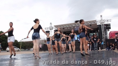 Grupo jovem realiza apresentação de dança sobre piso escuro em espaço aberto.