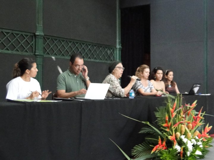 Participantes compõem uma mesa de trabalho do XII Fórum Nacional de Dança diante de arranjo floral.